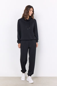 A woman wearing a black high-neck sweatshirt with ties and matching black pants, standing against a neutral background.
