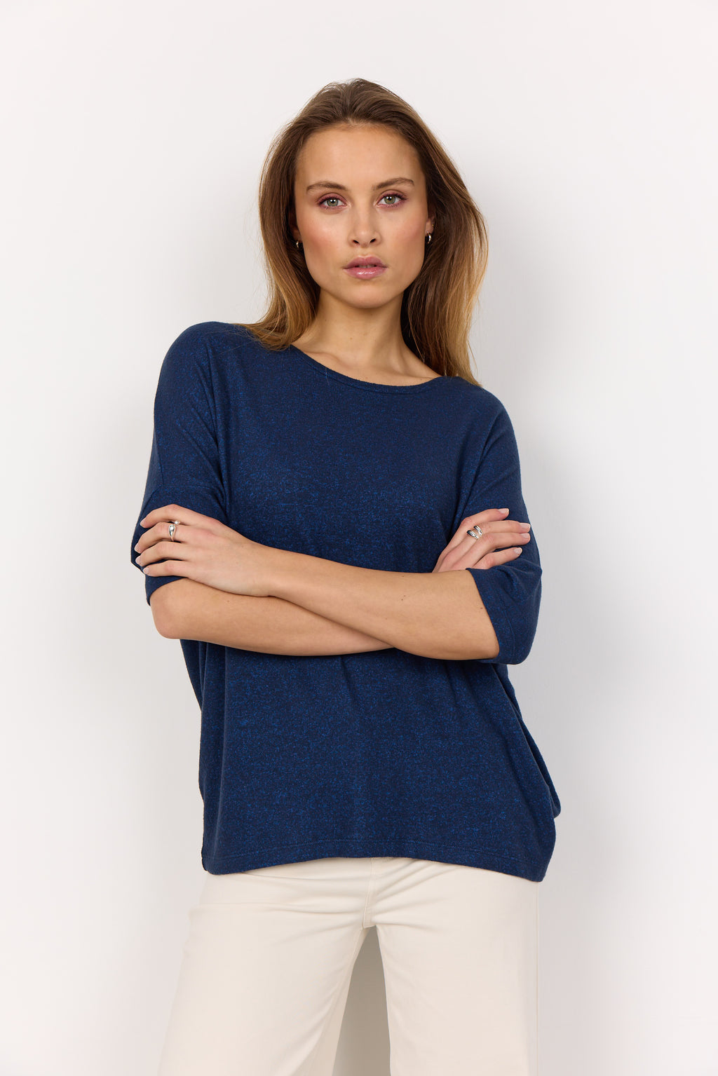 Woman wearing a blue sweater and white pants against a plain background. Sweater has a relaxed fit, 3/4 sleeves, and round neckline. 