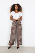 Load image into Gallery viewer, Woman wearing a white top and brown patterned pants against a white background
