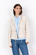 Load image into Gallery viewer, Woman wearing a  cream hooded windbreaker jacket over a light blue shirt and blue jeans on a white background
