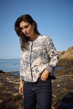 Load image into Gallery viewer, Woman wearing a navy floral bomber jacket standing on a rocky beach with ocean and clear blue sky in the background.
