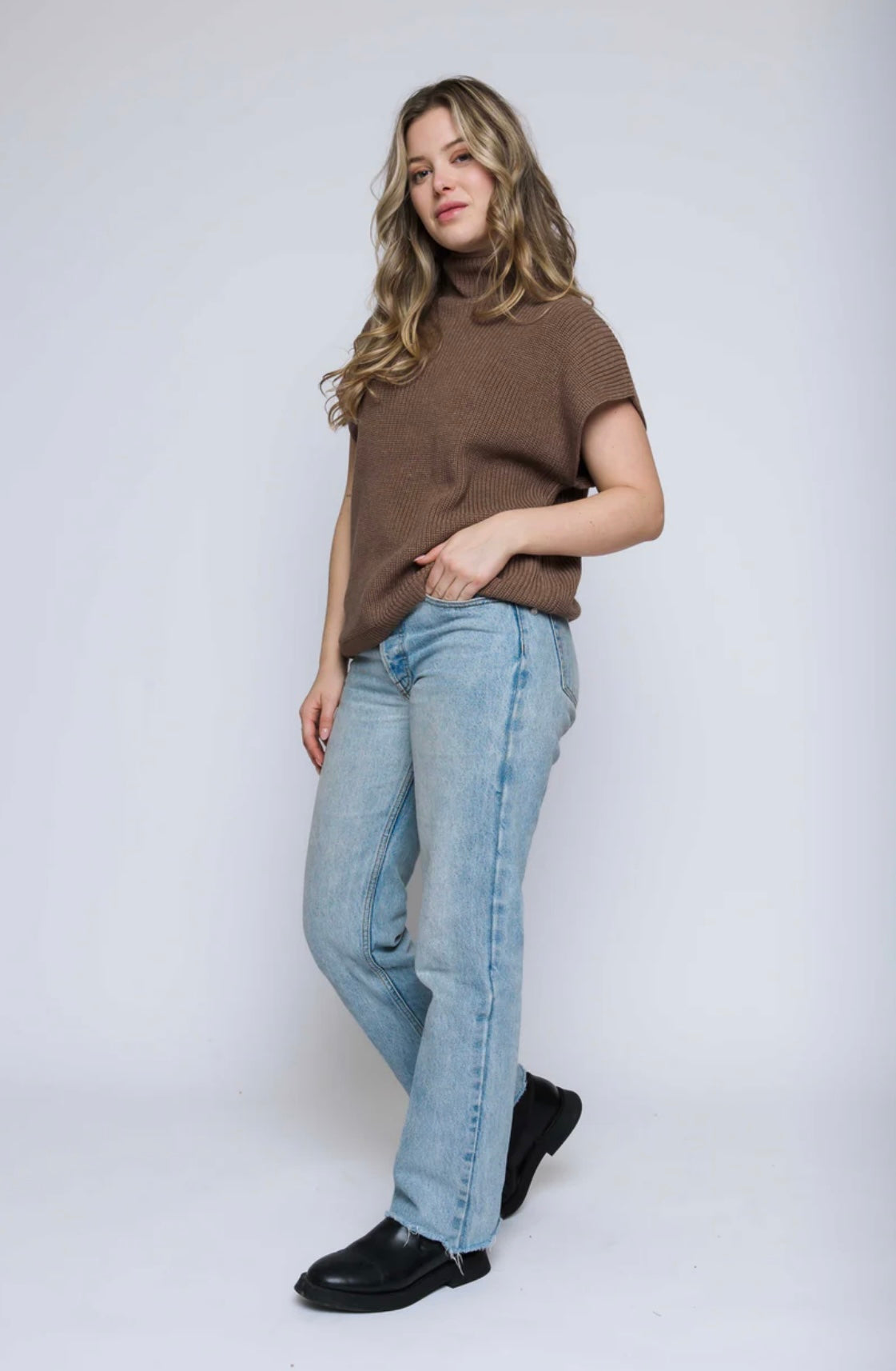 Woman wearing a brown turtleneck sweater vest and blue jeans on a white background