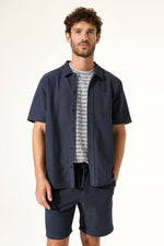 Load image into Gallery viewer, Man wearing a navy short-sleeve shirt over a patterned shirt with navy shorts on a white background
