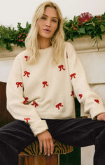 Load image into Gallery viewer, Woman wearing a cream sweater with red bow patterns sitting in front of a decorated fireplace.
