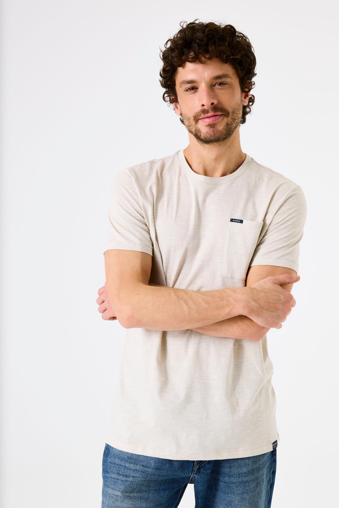 Man wearing a beige t-shirt with a brand logo on breast pocket on a white background