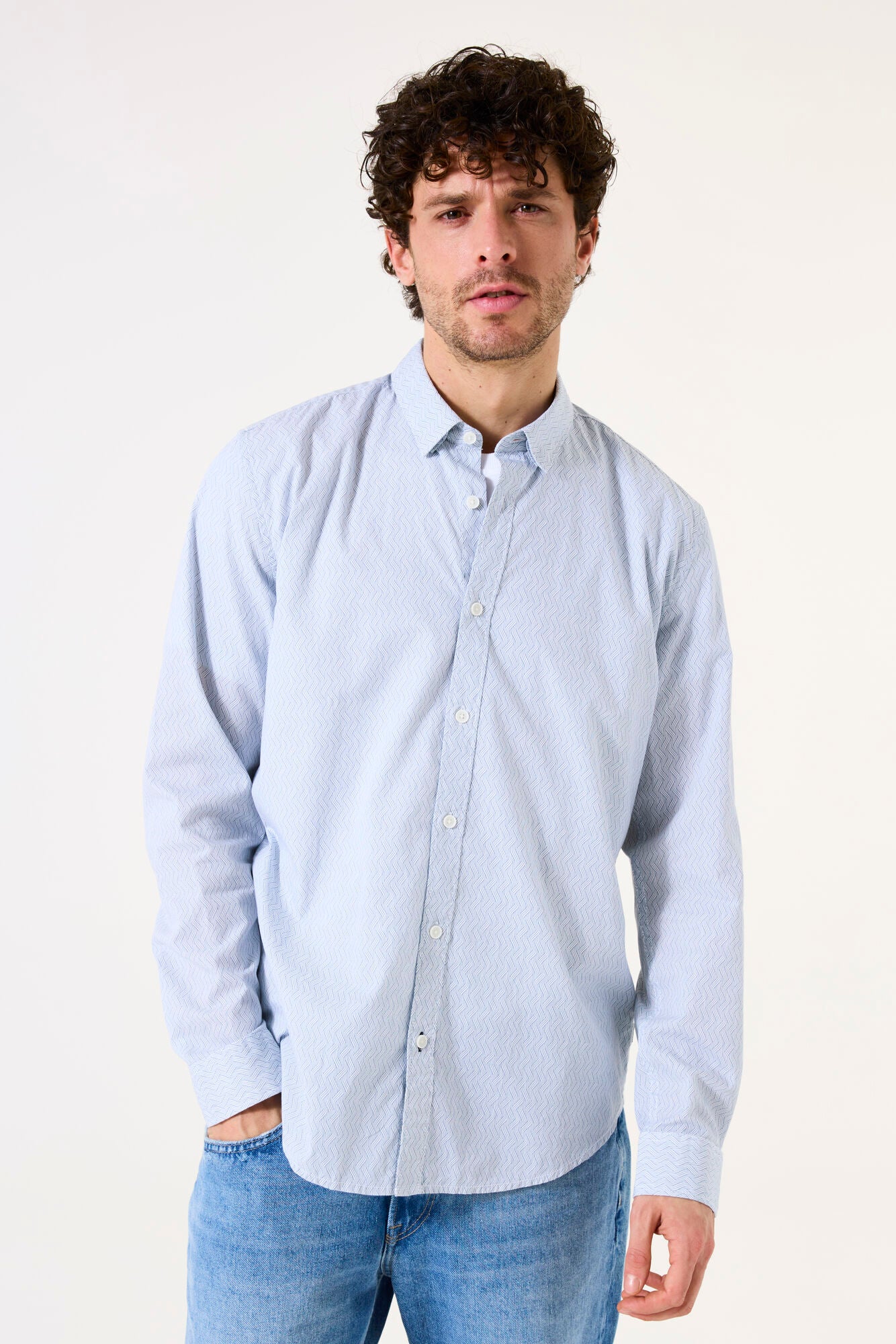 Man wearing a light blue and white button-up shirt and jeans against a plain background